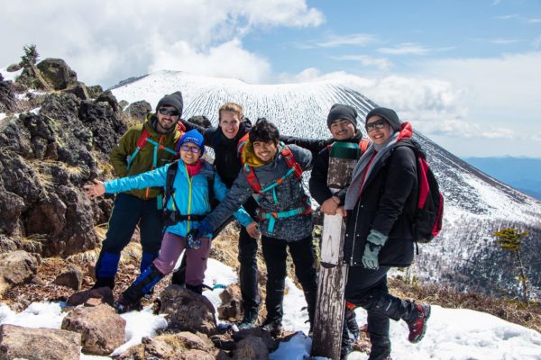 Covid-19 Has Strengthened My Character : Lhamo Hiking at Mount Asama with a nice group and outdoor education teacher. The group is spreading their arms and embracing on a ridge with the snow capped mountain in the mid ground. Blue mountains transition into a blue and cloudy sky in the background