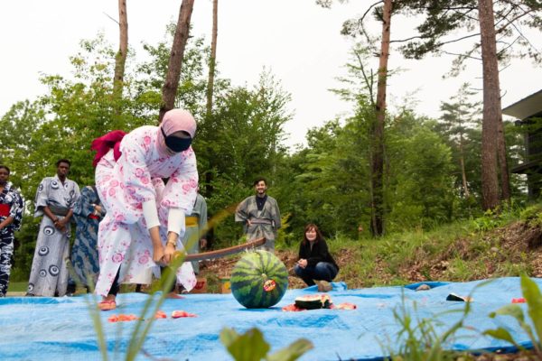 Blind folded girl in a pink yukata and pink hijab attempting to smash a watermelon sitting on a blue tarp with a wooden kendo sword for a japanese natsu matsuri or summer festival