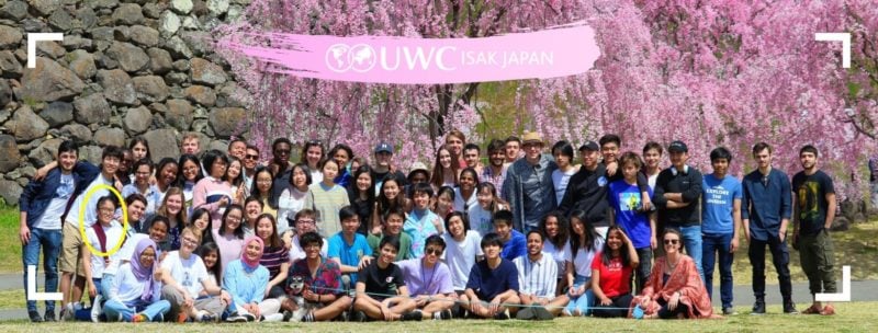 Covid-19 Has Strengthened My Character : Lhamo Sits With Her Whole graduation class , who are seated in four rows, everyone smiles and in the mid day sun. In the background to the left is a rock retaining wall at Ueda castle, to the right are breathtaking pink sakura trees in full bloom