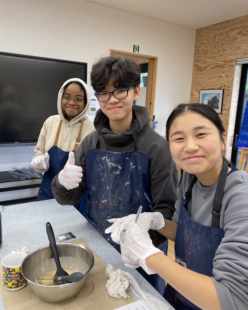 Three UWC ISAK Japan students smiling while working on their EcoSoap project during Fall Project Week 2024. They are wearing aprons and gloves as they mix soap ingredients in a bowl.