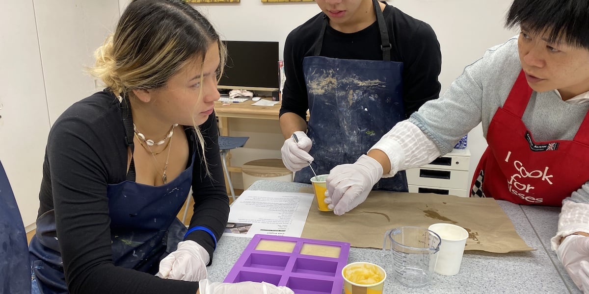 A UWC ISAK Japan student listens intently while being instructed by a mentor during the EcoSoap project. Two other students in aprons and gloves work on pouring soap mixtures into molds during Fall Project Week 2024.