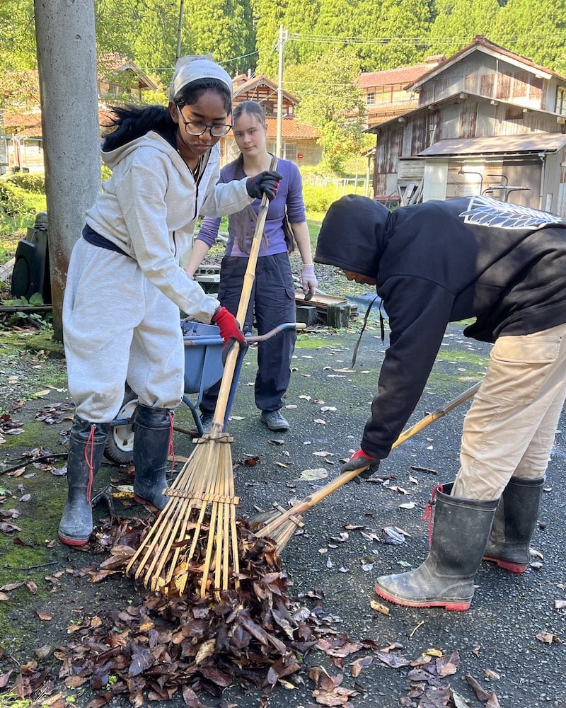 UWC ISAK Japan students working together to rake and collect leaves during their Nagano Harvest Project as part of Fall Project Week 2024. They are dressed in work clothes and boots, engaging in outdoor activities in a rural setting.