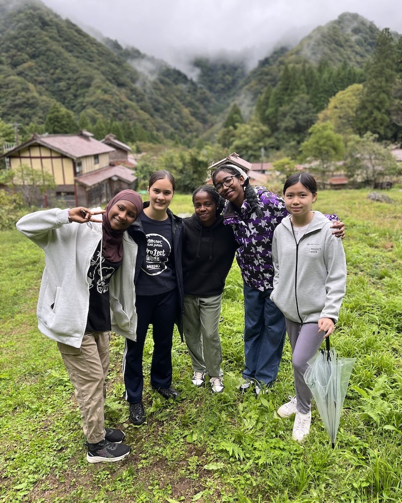 A group of UWC ISAK Japan students smiling and posing together during the Nagano Harvest Project in Fall Project Week 2024. They are standing in a lush, green rural setting with misty mountains in the background.