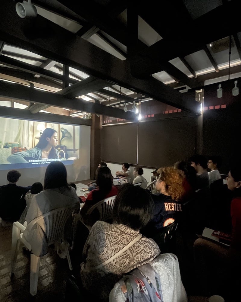 Audience members sit attentively in a dimly lit room, watching a film projected on a large screen. The film features a young woman in a close-up shot with subtitles. The cozy setup includes plastic chairs and informal seating arrangements, creating an intimate atmosphere for the film screening.