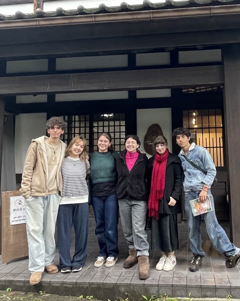 A group of six young people standing in front of a traditional Japanese building. They are casually dressed and smiling at the camera, with a cozy and friendly atmosphere. The wooden structure of the building adds a rustic charm, and the group appears to be part of a collaborative or creative project.