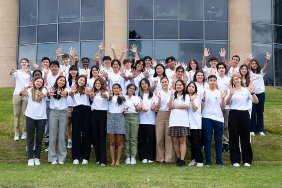 A vibrant group photo of students from different parts of the world who came together for the GCI Summit. They stand on a grassy lawn, smiling brightly and raising their hands in celebration, with a modern glass building in the background symbolizing collaboration and global connection