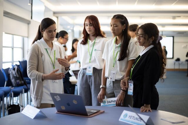 A group of students engaged in an interactive discussion around a laptop during a collaborative workshop at the GCI Summit. The presenter gestures confidently, explaining a concept, while the others listen attentively.