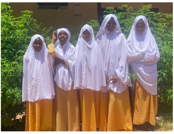 Five young girls are supported by the Stars Under the Refuge project. They are dressed in uniforms, consisting of white hijabs and golden-yellow dresses. The bright sunlight highlights their smiles and expressions as they pose together. 