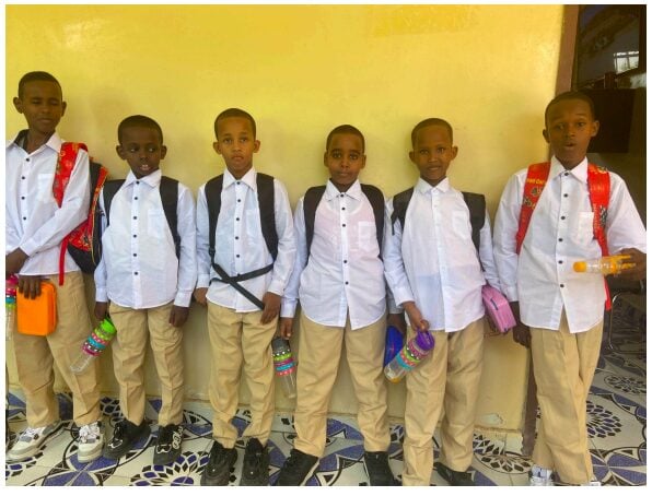 Six young boys supported by the Stars Under the Refuge project. They are dressed in matching uniforms, consisting of white button-up shirts, beige pants, and black or white shoes. Each boy carries a backpack and holds a colorful water bottle or item in their hands. 