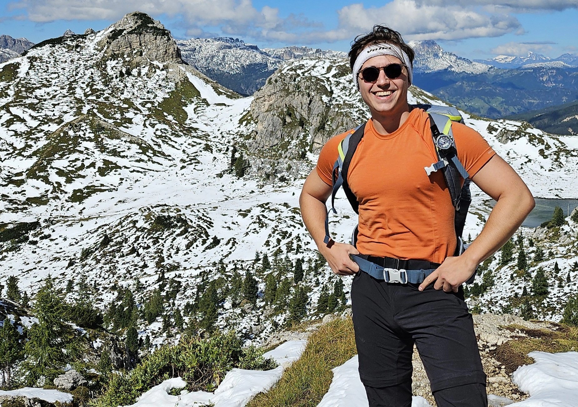 Viktor, wearing an orange T-shirt, sunglasses, and a headband standing on a snowy mountainous landscape. He has a backpack and is surrounded by rocky terrain with patches of snow and greenery under a bright blue sky with scattered clouds.