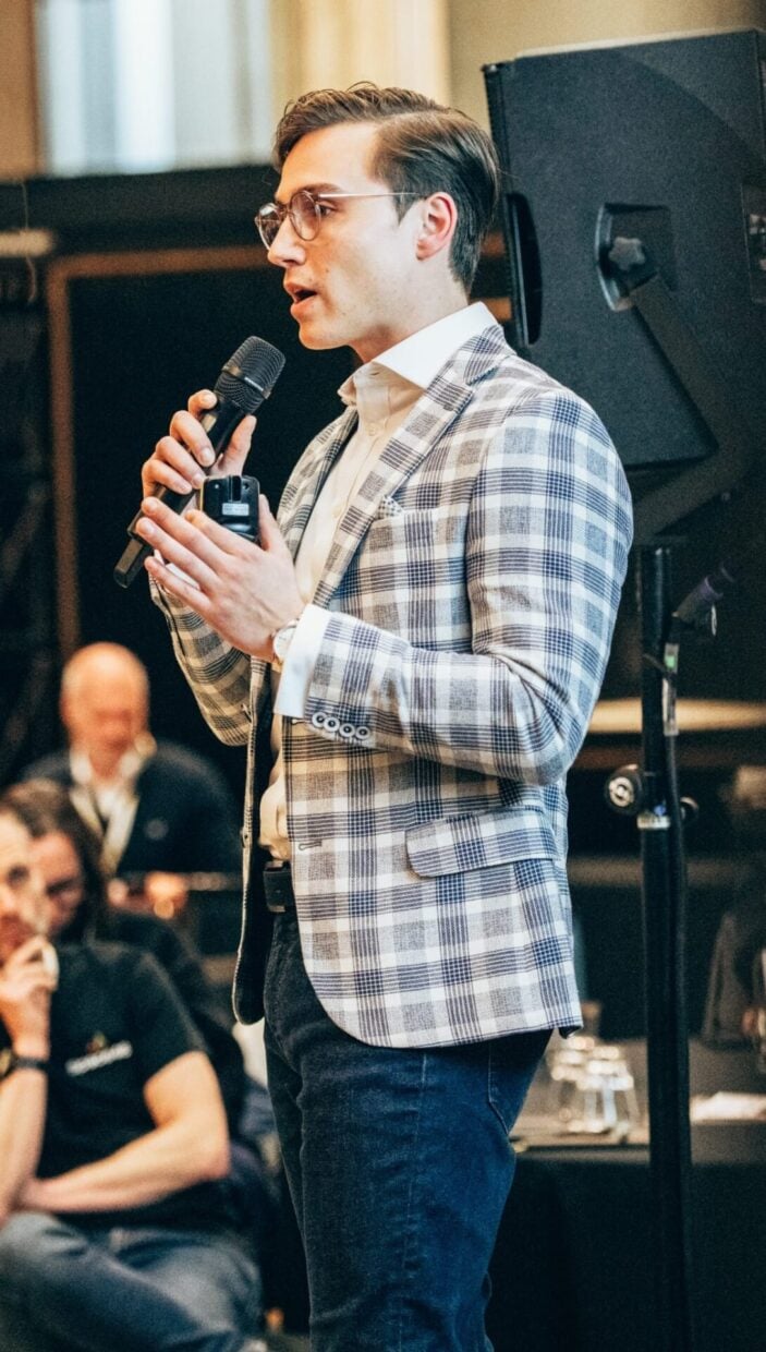 Viktor is speaking at a professional event, holding a microphone in one hand and gesturing slightly with the other. He is wearing a plaid blazer, a white shirt, and jeans, with an audience visible in the background