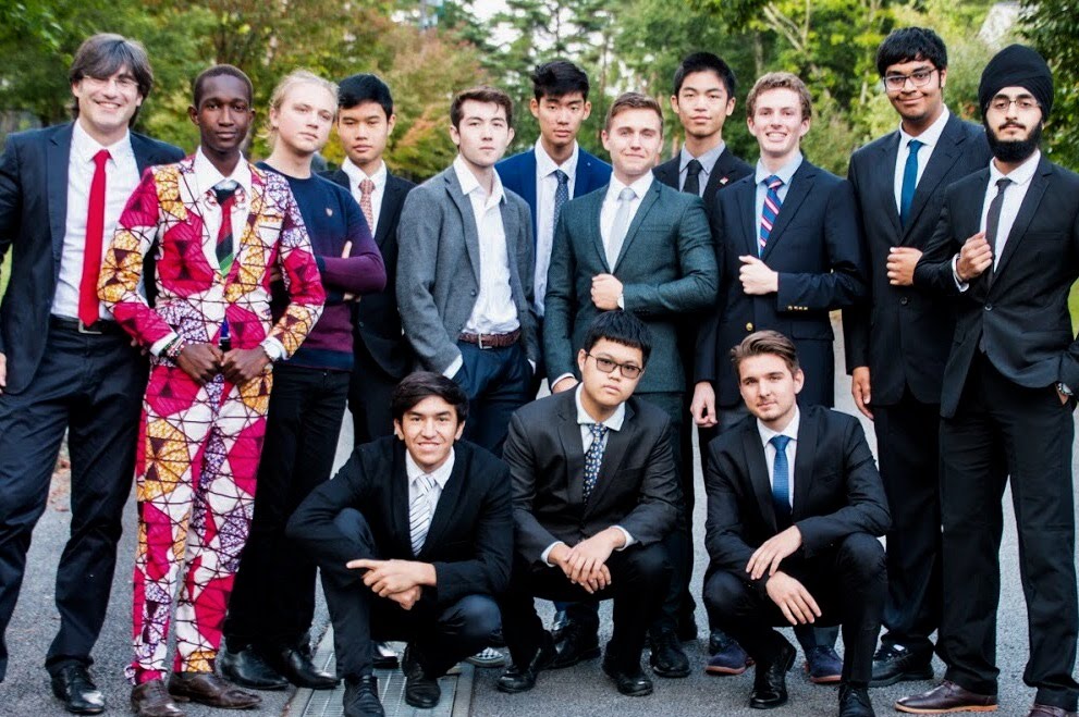 A group of 13 ISAK students posing together outdoors, dressed in formal suits. One individual stands out wearing a vibrant, patterned red, pink, and white suit. They are all smiling or looking confidently at the camera, with greenery and a pathway in the background. They have since graduated.