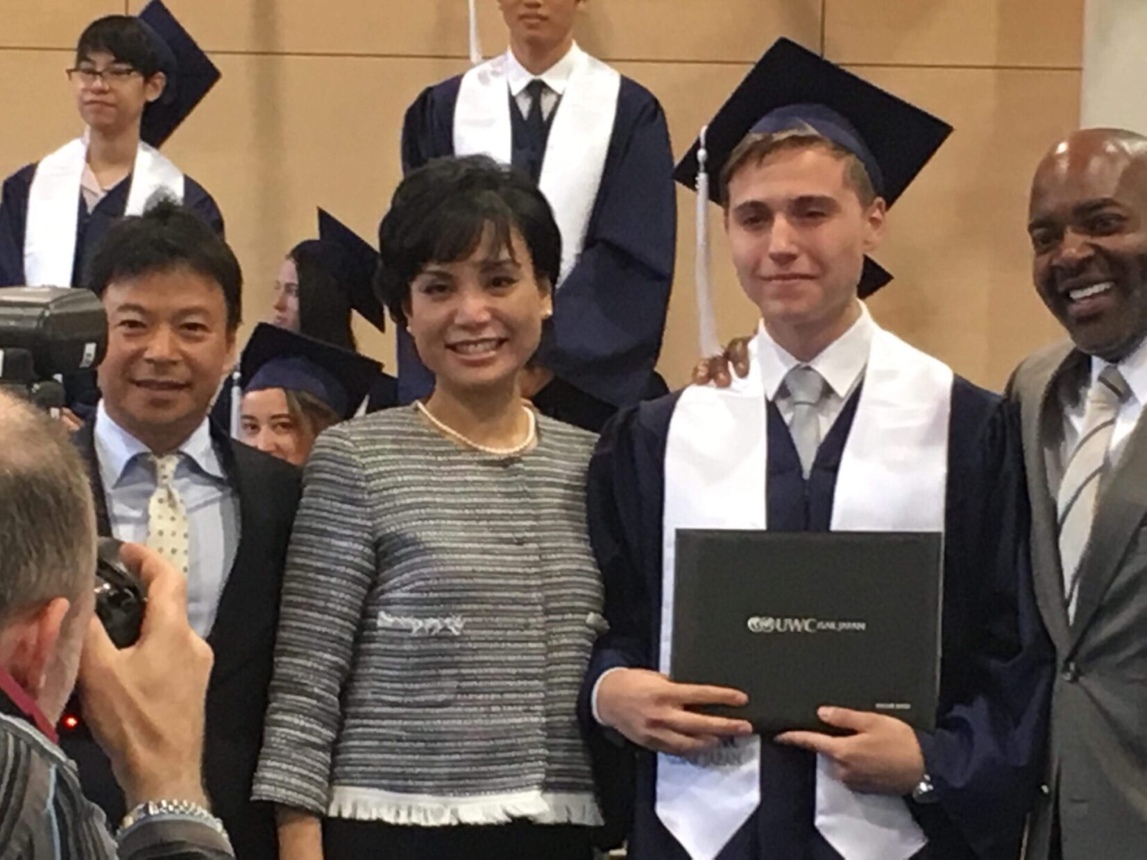 Viktor, dressed in a navy blue graduation cap and gown with a white stole, holds a diploma during the ISAK Japan graduation ceremony. He stands with Lin Kobayashi, who is smiling and wearing a tweed jacket with pearls, alongside two other individuals in formal attire. Other graduates in similar attire can be seen in the background, while a photographer captures the moment in the foreground.